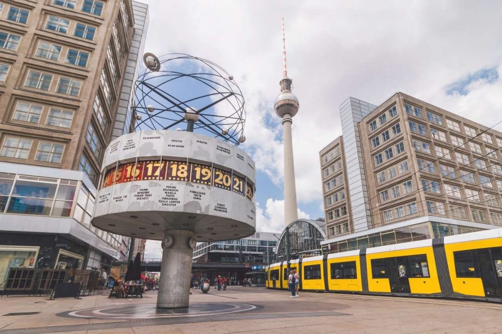 Weltzeituhr und Fernsehturm auf dem Alexanderplatz in Berlin mit vorbeifahrender gelber Straßenbahn und umliegenden Gebäuden.