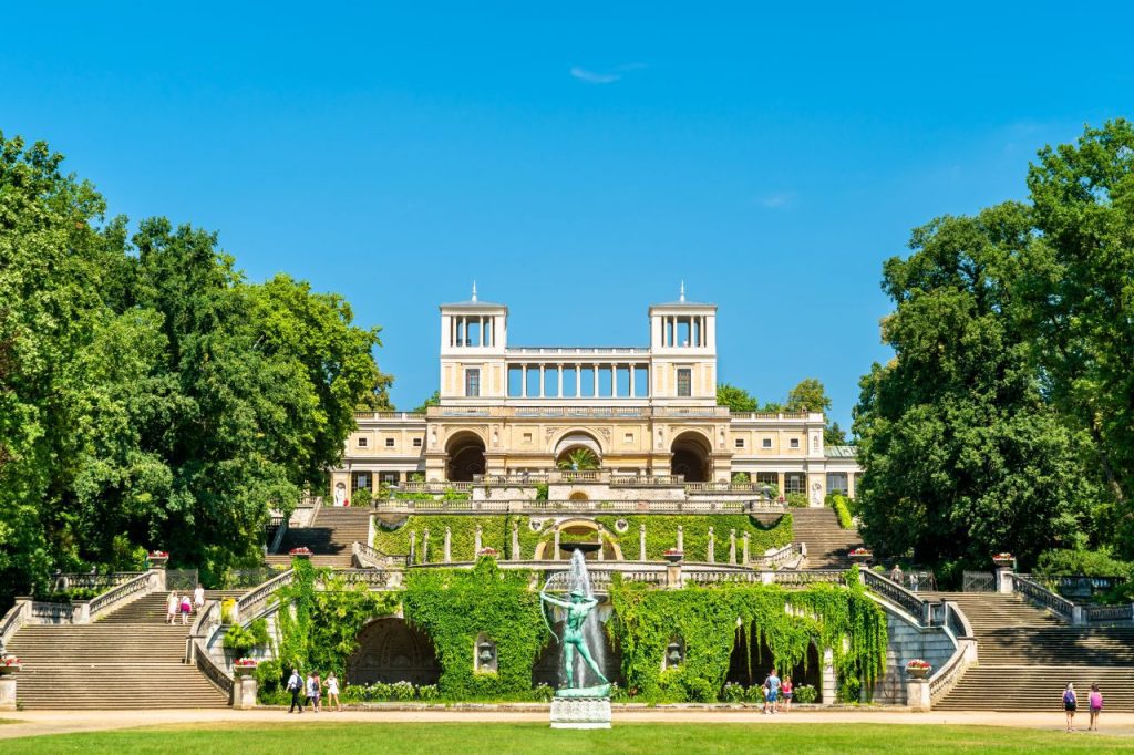 Italienische Renaissancearchitektur des Orangerieschlosses im Park Sanssouci in Potsdam mit Terrassen, Treppen und Brunnenstatue bei blauem Himmel.