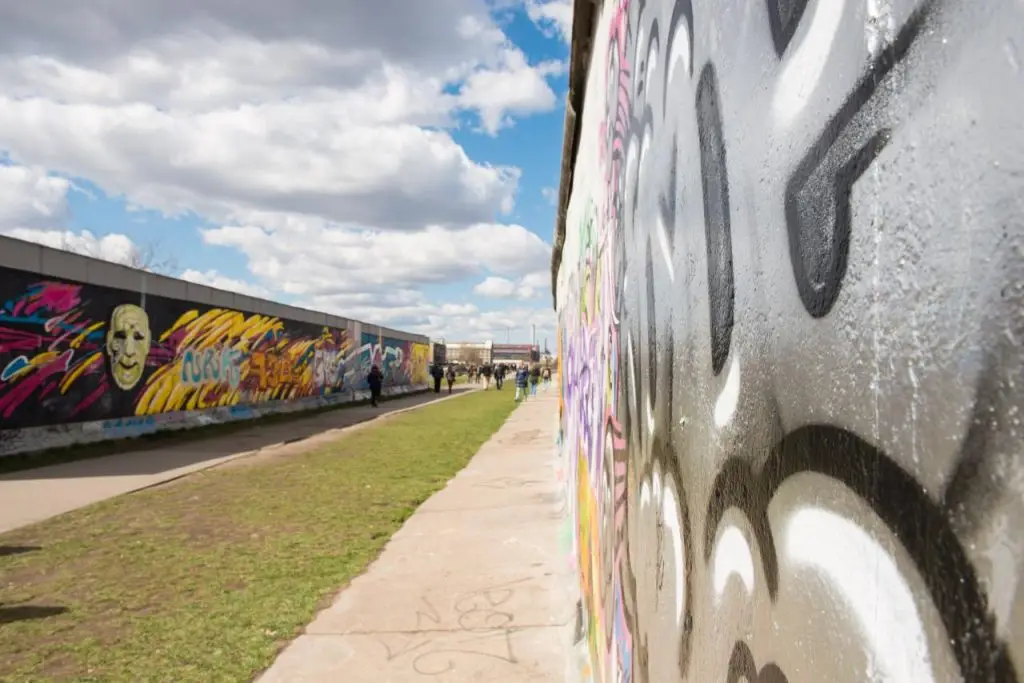 Bemalte Berliner Mauer an der East Side Gallery mit Graffiti und Spaziergängern bei wolkigem Himmel.