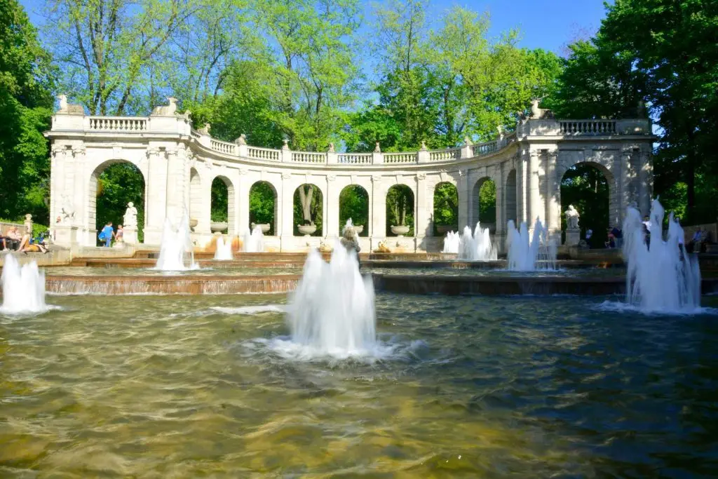 Neptunbrunnen im Volkspark Friedrichshain in Berlin mit Wasserfontänen und halbkreisförmiger Kolonnade bei Sonnenschein.